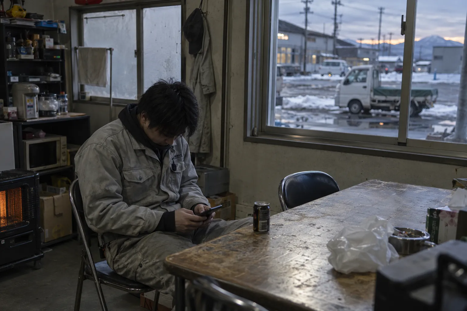 冬の夕方、工場の休憩所でスマートフォンを見る作業着の若い男性